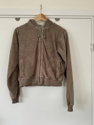 Brun velour hoodie med dragkedja - storlek S - Mjuk och len brun huvtröja / hoodie från Emilia i storlek S. Klassisk modell med dragkedja och fickor framtill. Tröjan har en huva med dragsko och ribbade muddar i ärmslut och nederkant. 80% bomull, så bra material som andas. Fint skick!