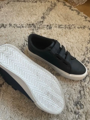 Svarta sneakers med kardborreband - Snygga svarta sneakers med tre kardborreband och vit sula. Skorna har en klassisk låg modell och är tillverkade i ett slätt syntetmaterial. Perfekta för dig som gillar enkel och stilren design.OBS! ÄR ANVÄNDA 1 gång men är i bra skick annars 
