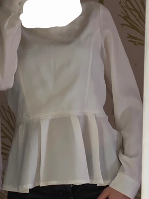 Vit peplumblus med långa ärmar - Snygg vit blus med peplumskärning och långa ärmar. Blusen har en rund halsringning och är tillverkad i ett lätt, något transparent tyg som ger en luftig känsla. Perfekt att styla med jeans eller kjol för en trendig look.