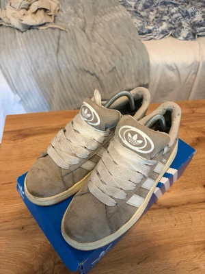 Grå Adidas sneakers med vita detaljer - Snygga grå sneakers från Adidas med klassiska vita ränder på sidorna och stor vit logga på plösen. Skorna har breda vita skosnören och en låg siluett. Ovandelen är i mocka och textil, perfekt för en avslappnad streetstil.