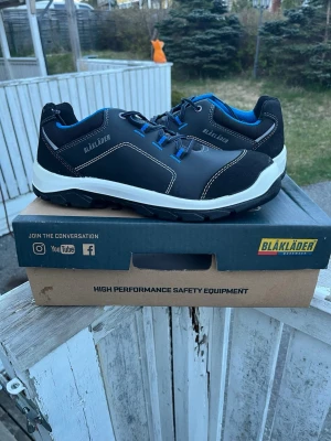 Svarta sneakers från Blåkläder - Svarta sneakers från Blåkläder med blå detaljer och vit sula. Skorna har tåhätta, vattenavvisande ovandel och slitstark yttersula i PU/gummi. Insidan är fodrad med blå textil och skon har snörning framtill. Perfekt för dig som gillar en sportig och robust stil.                                                      Orginal pris 1500kr