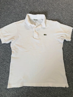 Vit pikétröja från Lacoste - Klassisk vit pikétröja från Lacoste med broderad grön krokodil på bröstet. Den är ca 65cm på längden och ca 51cm på brädden. Hör av dig vid frågor och funderingar!🤗
