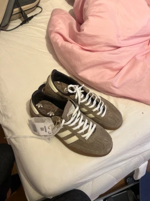 Adidas beige sneakers med vita ränder - HELT NYA!!!Snygga Adidas sneakers i beige mocka med klassiska vita ränder på sidorna och vit snörning. Yttersulan är i gummi med retroinspirerat mönster. Perfekta för dig som gillar stilrena och tidlösa sneakers med streetkänsla.