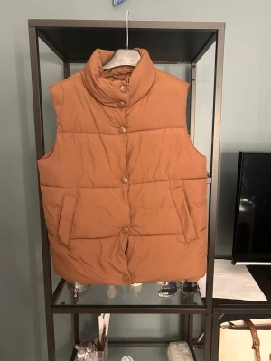 Beige quiltad dunväst med hög krage - Snygg beige dunväst med hög krage och tryckknappar framtill. Västen har två stora fickor på sidorna och en quiltad design som ger en puffig look. Perfekt för lager på lager och enkel att styla till olika outfits.
