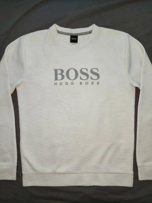 Vit sweatshirt från Hugo Boss - Vit sweatshirt från Hugo Boss med stor grå logotyp på bröstet. Tröjan har rund halsringning, långa ärmar och ribbade muddar vid ärmslut och nederkant. Tillverkad i ett mjukt och strukturerat material som känns skönt mot huden.