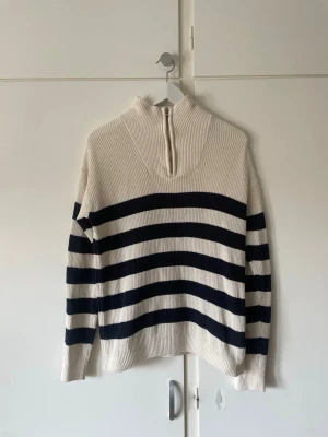 Randig stickad tröja från H&M - Stickad tröja från H&M i vitt med breda mörkblå ränder. Tröjan har hög krage med dragkedja och är ribbstickad för en snygg struktur. Perfekt för dig som gillar en avslappnad men trendig look.