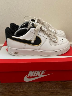 Nike Air Force 1 vit/svart/guld - Nike Air Force 1 sneakers i vitt läder med svart Swoosh och guldkant. Klassisk låg modell med perforerad tå, vita skosnören och svart vadderad häl. Snygg kontrast mellan färgerna och ikonisk Nike-logga på plös och häl.