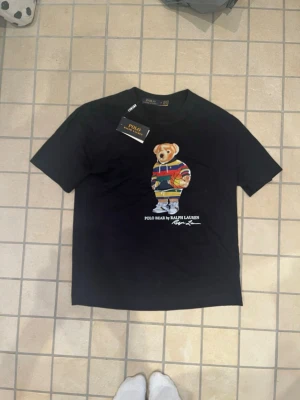 Svart Polo Ralph Lauren t-shirt med björn - Svart t-shirt från Polo Ralph Lauren med klassisk björn-tryck på bröstet. T-shirten har rund halsringning och korta ärmar. Björnen bär en färgglad hoodie och håller en basketboll. Perfekt för dig som gillar streetstyle och ikoniska prints.