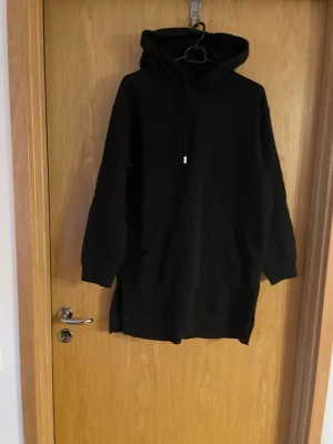 Svart hoodieklänning med huva - En svart hoodieklänning med huva och dragsko. Modellen har långa ärmar, stor ficka framtill och är tillverkad i mjukt sweatshirtmaterial. Perfekt för en avslappnad och chill stil.
