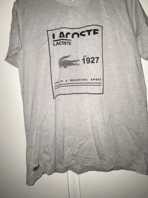 Grå Lacoste t-shirt med tryck - Snygg grå t-shirt från Lacoste med stort svart tryck framtill och klassisk broderad krokodil nertill. T-shirten har rund hals och korta ärmar, perfekt för en avslappnad och stilren look.