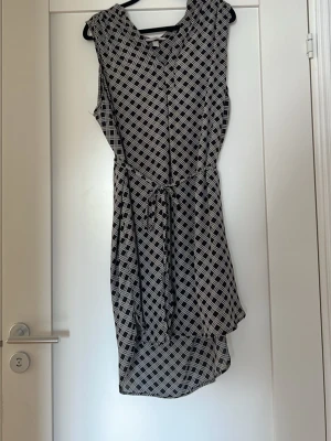 Svartvit mönstrad ärmlös klänning - Snygg ärmlös klänning i svartvitt rutmönster med knyt i midjan. Klänningen har en avslappnad passform och är tillverkad i ett lätt material som passar perfekt för varmare dagar. Den har en kort längd och ett stilrent, modernt uttryck.