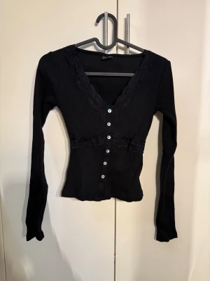 Svart ribbad topp med spets och knappar - Snygg svart ribbad topp med långa ärmar, v-ringning och dekorativa spetsdetaljer vid halsringningen och midjan. Framtill sitter vita knappar hela vägen ner. Figurnära passform som framhäver siluetten, perfekt till jeans eller kjol.