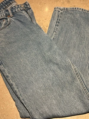 Blå raka jeans i klassisk modell - Säljer ett par blå jeans i klassisk rak modell med hög midja och fem fickor. Jeansen har en ljus tvätt och är tillverkade i 100% bomull. Perfekta för en avslappnad och tidlös stil.