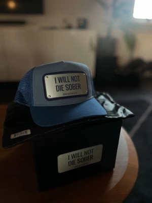 Blå keps John Hatter & Co - Snygg blå truckerkeps från John Hatter & Co med metallskylt framtill där det står 'I WILL NOT DIE SOBER'. Kepsen har justerbar snapback och mesh på sidorna och bak för extra ventilation. Perfekt statement piece för dig som vill sticka ut. 