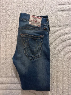 True Religion Rocco relaxed skinny jeans - Säljer ett par blå True Religion Rocco relaxed skinny jeans med klassisk kontrastsöm och ikonisk bakficka med broderad logga. Jeansen har orange detaljer och är tillverkade i bomull. Perfekta för dig som gillar streetwear och vill ha en avslappnad men slim passform.