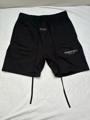 Svarta Essentials shorts med snörning - Svarta shorts från Fear of God Essentials med långa snören i midjan och tryckt logga på vänster ben. Modellen har elastisk midja, sidofickor och en bakficka. Perfekta för en avslappnad och trendig look.