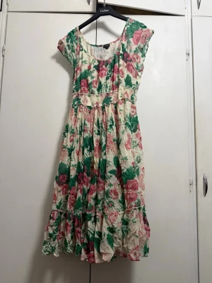 Blommig sommarklänning med volang - Söt blommig klänning i vitt med rosa och gröna detaljer. Klänningen har korta ärmar, markerad midja med smock och volangkant nertill. Perfekt för varma dagar och har en luftig, romantisk stil.