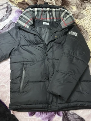 Svart pufferjacka från Burberry - Snygg svart pufferjacka från Burberry med klassiskt rutigt foder i grått, rött och vitt. Jackan har dragkedja och knappar framtill, två sidofickor med dragkedja och logga på ärmen. Perfekt för kalla vinterdagar.