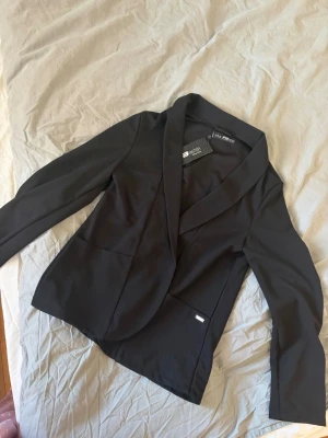 Svart kavaj från Las Pepas - Stilren svart kavaj från Las Pepas med klassisk krage och två framfickor. Kavajen har en figurnära passform och är tillverkad i ett mjukt, slätt material. Perfekt för en trendig och uppklädd look. LITEN I STORLEK 