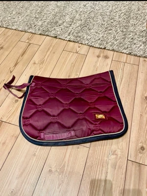 Vinrött schabrak från Eqestrian Stockholm - Säljer ett vinrött schabrak från Equestrian Stockholm  med svart och guldig kant. Schabraket har quiltat mönster och en liten guldfärgad logga på sidan. Perfekt för dig som vill ha stil på ridturen.