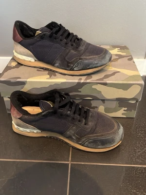 Svarta sneakers från Valentino - Snygga svarta sneakers från Valentino med detaljer i vinrött och grått. Skorna har en mix av mocka och mesh, svarta skosnören och beige sula. Perfekta för dig som gillar streetstyle och exklusiva märken.
