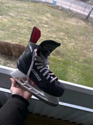 Svarta Bauer hockeyskridskor - Säljer ett par svarta hockeyskridskor från Bauer med vita snören och detaljer i rött och vitt. Skridskorna har en robust plastkonstruktion och klassisk form, perfekta för isträning eller match. Märket syns tydligt på sidan och plösen.