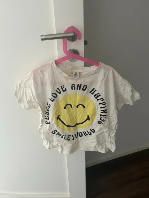 Vit SmileyWorld t-shirt med tryck - Vit t-shirt från SmileyWorld med stort gult smileytryck och texten 'Peace Love and Happiness' på framsidan. Kortärmad och croppad modell i mjukt bomullstyg. Perfekt för en avslappnad och lekfull stil.