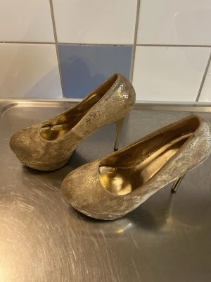 Guldiga pumps med ormmönster - Säljer ett par glittriga guldiga pumps från Gemma i storlek 39. Skorna har ett snyggt ormmönster, rundad tå och höga smala klackar. Insidan är också guldfärgad vilket ger en extra lyxig känsla. Perfekta för dig som vill sticka ut!