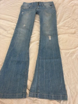 Ljusblå bootcut jeans med slitningar - Snygga ljusblå bootcut jeans med slitna detaljer på låren och knäna. Jeansen har klassisk femficksmodell, låg midja och vida ben. Perfekta för en avslappnad och trendig look.