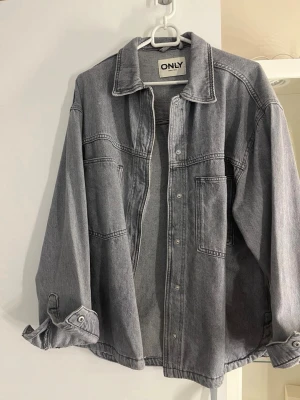 Grå jeansjacka från ONLY - Snygg grå jeansjacka från ONLY med klassisk krage, knappstängning framtill och två stora bröstfickor. Jackan har avslappnad passform och nedhasade axlar för en cool och trendig look. Perfekt att slänga över en hoodie eller t-shirt.