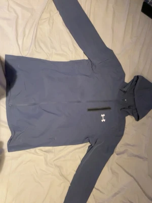 Blå vindjacka från Under Armour - Snygg blå vindjacka från Under Armour med huva och dragkedja framtill. Jackan har en bröstficka med dragkedja och logga på bröstet. Perfekt för blåsiga dagar och sportiga outfits.OBS Dem har 2 små hål på högerarmen man märker dem knappt originellt köpt för 1200kr