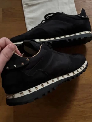 Svarta sneakers med nitar från Valentino - Snygga svarta sneakers från Valentino med coola silverfärgade nitar runt sulan. Skorna har en mix av mocka och textil, svart yttersula med grovt mönster och beige innerfoder. Perfekta för dig som vill sticka ut med en edgy detalj.