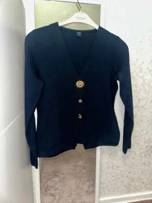 Marinblå kofta från Kiabi med guldknappar - Stilren marinblå kofta från Kiabi med djup v-ringning och tre dekorativa guldknappar, varav en är formad som en blomma. Långärmad och i mjukt stickat material, perfekt för att lyfta din outfit.