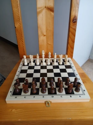 Schackspel i trä med bräde - Säljer ett klassiskt schackspel med bräde i trä. Pjäserna är i två färger, mörkbrunt och ljust trä, och brädet har svartvita rutor med bokstäver och siffror längs kanterna. Brädet har ett litet metallspänne för att hålla ihop det vid transport.