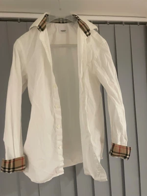 Vit skjorta från Burberry med rutiga detaljer - Vit skjorta från Burberry med klassiska rutiga detaljer på krage och ärmslut. Skjortan har knappar framtill och en tidlös design som passar perfekt till många olika stilar. Tillverkad i bomull för en skön känsla.