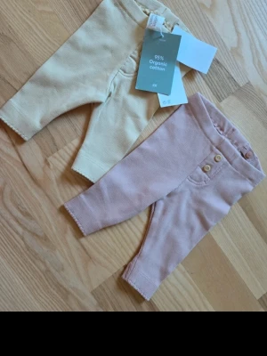 2-pack leggings H&M, beige & rosa - Två par mjuka leggings från H&M i storlek 56, ett par i ljusbeige och ett par i ljusrosa. Tillverkade i 95% ekologisk bomull med stretch. Båda har resår i midjan och det rosa paret har dekorativa knappar framtill. Perfekta för en chill och bekväm stil.