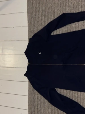 Mörkblå hoodie från Polo Ralph Lauren - Mörkblå hoodie med dragkedja från Polo Ralph Lauren. Klassisk design med vit broderad logga på bröstet och två fickor framtill. Perfekt för en avslappnad och stilren look.