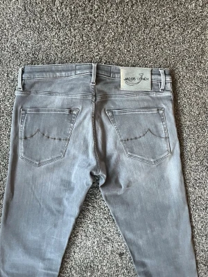 Gråa slim fit jeans från Jacob Cohen - Säljer ett par snygga gråa jeans från Jacob Cohen med slim fit passform. Jeansen har klassiska bakfickor med diskret söm och ett ljust patch med logga bak i midjan. Perfekta för dig som gillar stilrena och moderna jeans. Storleken är 32, men tycker de passar mindre o sitter nu som 30/32.