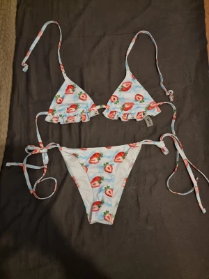 Bikini med volang och knyt - Söt bikini med ljusblå botten och jordgubbsmönster. Överdelen har volangkant och trekantsform med knyt i nacke och rygg. Underdelen har knyt i sidorna för justerbar passform. Perfekt för sommaren och strandhäng! ⚠️HELT OANVÄND⚠️