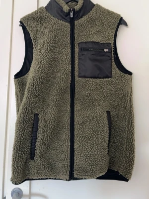 Olivgrön teddyväst med dragkedja - Säljer en olivgrön teddyväst med hel dragkedja framtill och svarta detaljer. Västen har en bröstficka i nylon med dragkedja samt två sidofickor. Materialet är mjukt och fluffigt, perfekt för lager på lager. Passar dig som gillar streetwear och avslappnad stil.