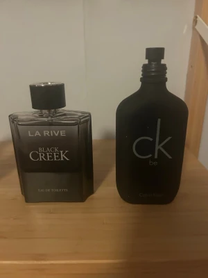 La Rive Black Creek & CK Be parfym - Två parfymer: La Rive Black Creek i en fyrkantig, mörkgrå glasflaska med svart lock och tydlig logga, samt Calvin Klein CK Be i en matt svart, rundad flaska med minimalistisk design. Båda flaskorna är stilrena och passar perfekt i badrumshyllan. La rive inte använd alls. Calvin Klein mycket använd. 