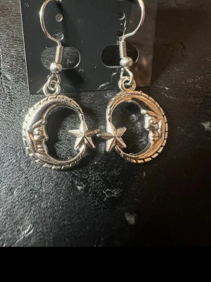 Silvriga örhängen med måne och stjärna - Unika silvriga örhängen i metall med hängande måne och stjärna. Månarna har ett ansikte i relief och stjärnorna sitter i mitten. Perfekta för dig som gillar mystiska och boho-inspirerade accessoarer.