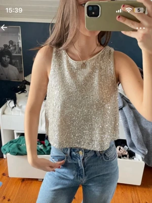 Glittrig croppad topp i silver - Säljer en glittrig croppad topp i silver med ärmlös design. Perfekt för dig som vill sticka ut och glänsa. Toppens material är syntet och den har en loose passning som gör den bekväm att bära.
