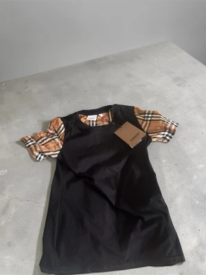 Svart topp med rutiga puffärmar Burberry - Säljer en svart topp från Burberry med klassiska beige och bruna rutiga puffärmar. Toppen har rund halsringning och är tillverkad i mjuk bomull. Perfekt statement-plagg för dig som gillar ikoniska detaljer och stilren design. skön t shirt.