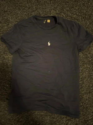 Svart t-shirt från Ralph Lauren - Klassisk svart t-shirt från Ralph Lauren med den ikoniska vita polospelaren broderad på bröstet. T-shirten har rund hals och korta ärmar, perfekt för en stilren och enkel look.