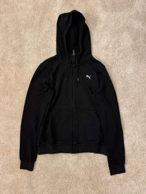 Svart hoodie från Puma med dragkedja - Svart hoodie från Puma med vit logga på bröstet, dragkedja framtill och två stora fickor. Modellen har huva med snören och ribbade muddar vid ärmslut och nederkant. Perfekt för en avslappnad och sportig look. Passar även S.