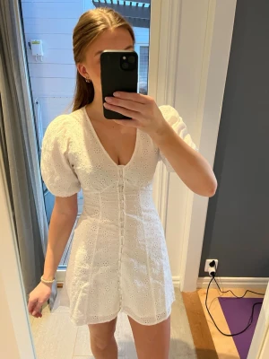 Klänning - Säljer en vit klänning med v-ringning, puffärmar och broderade blommönster. Perfekt för studenten eller som sommarklänning 