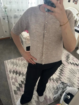 Beige kortärmad kofta med pärlknappar - Säljer en beige, kortärmad kofta med rund hals och pärlknappar framtill. Koftan är stickad i ett mjukt material och har en rak passform som ger en clean och stilren look. Perfekt att matcha med både jeans och kjol.