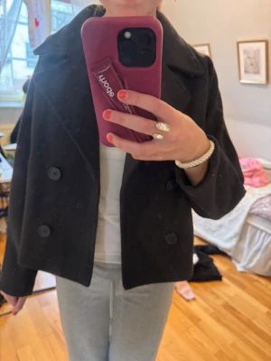 Svart dubbelknäppt kappa - Stilren svart kavaj med kappa och bred krage. Kortare modell som passar perfekt till både jeans och kjol. Klassisk design med stora svarta knappar och mjukt tyg. Är lite luddig och nopprig.