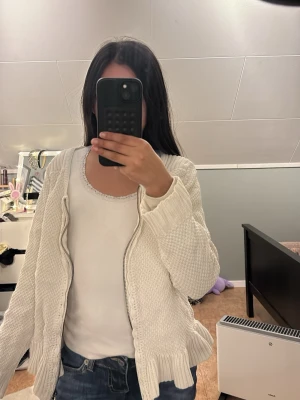 Vit stickad kofta med volangkant - Säljer en vit stickad kofta med dragkedja och volangdetaljer längs nederkanten. Koftan har långärmad design och strukturstickat mönster. Perfekt för att lägga över en topp eller t-shirt.